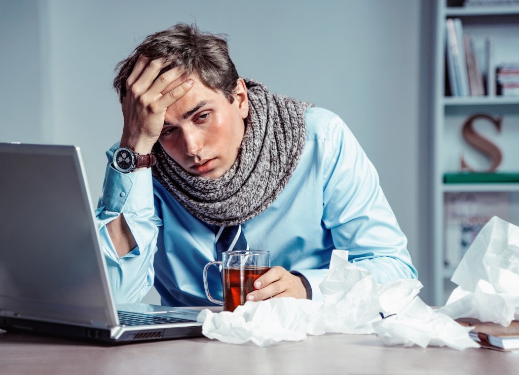 Flu symptoms include fever, chills, sore throat, dry cough, headache, muscle aches, runny or stuffy nose, and fatigue. Here, a man looks miserable and cold at his computer, while a bunch of beans sit nearby.