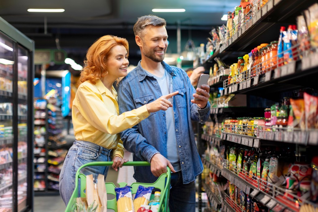 A European couple buys food in the supermarket, using a mobile phone in the aisle of the store